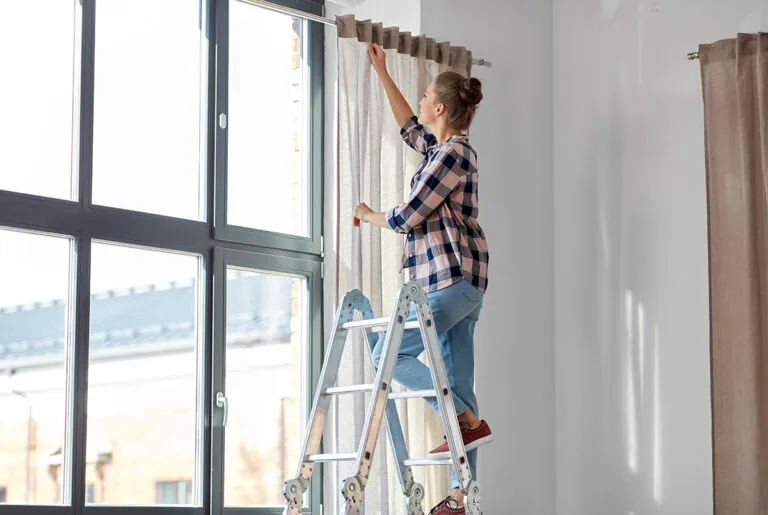 woman on ladder hanging curtains at home